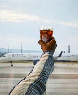 A person holding a Starbucks iced coffee in front of a Delta plane