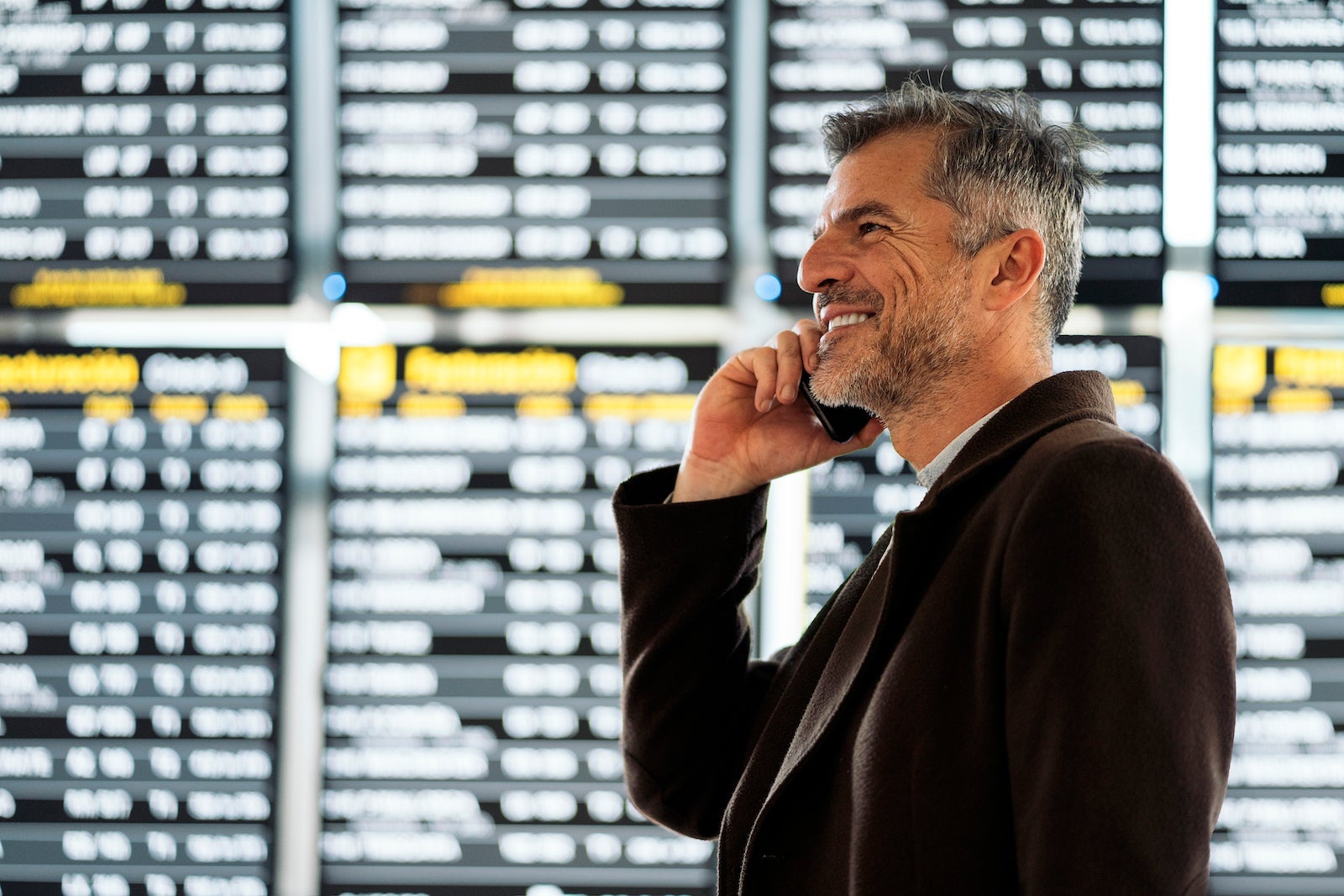 Photo of a side view of a caucasian business man talking on his phone in the airport. He is near the timetable of departures. He is smiling.