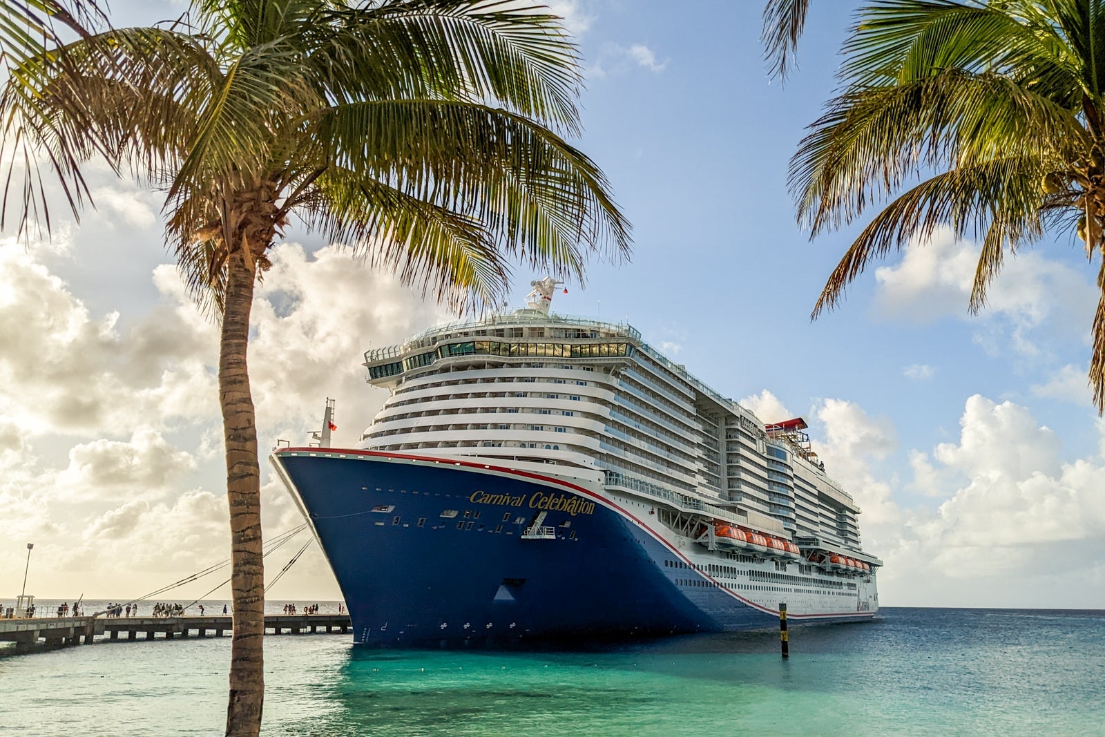 A white cruise ship with a blue hull docked in teal-green water a palm tree on either side and blue sky with white clouds in the background
