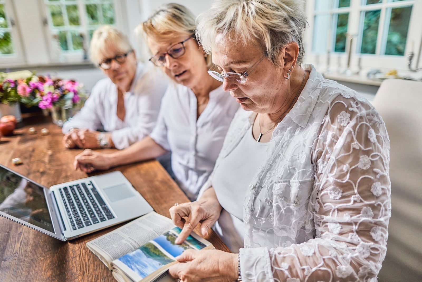 elderly friends look at a guide book and use a computer to plan a vacation