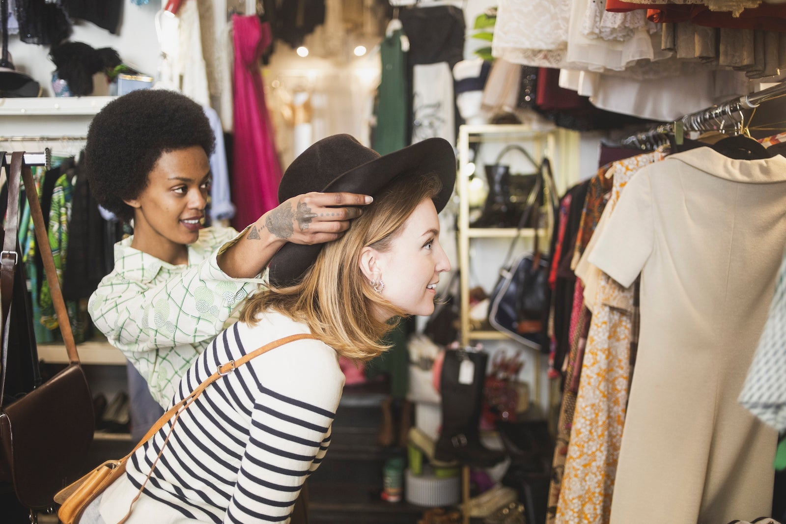 A store owner puts a hat on a customer's head while they look at a mirror together
