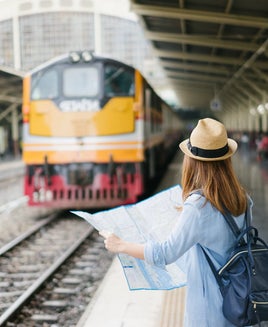 Young woman traveler with sky blue backpack and hat looking the map with train background at train station Bangkok. Traveling in Bangkok Thailand. Travel concept