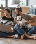 A young couple sits with a dog and moving boxes while shopping online with a laptop