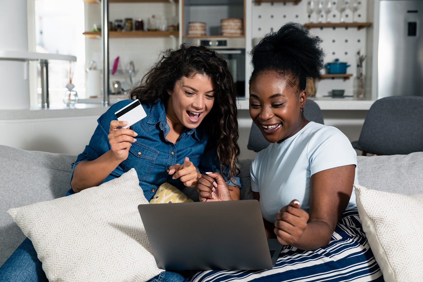 Two women sitting on a couch booking travel while looking at a laptop and holding a credit card
