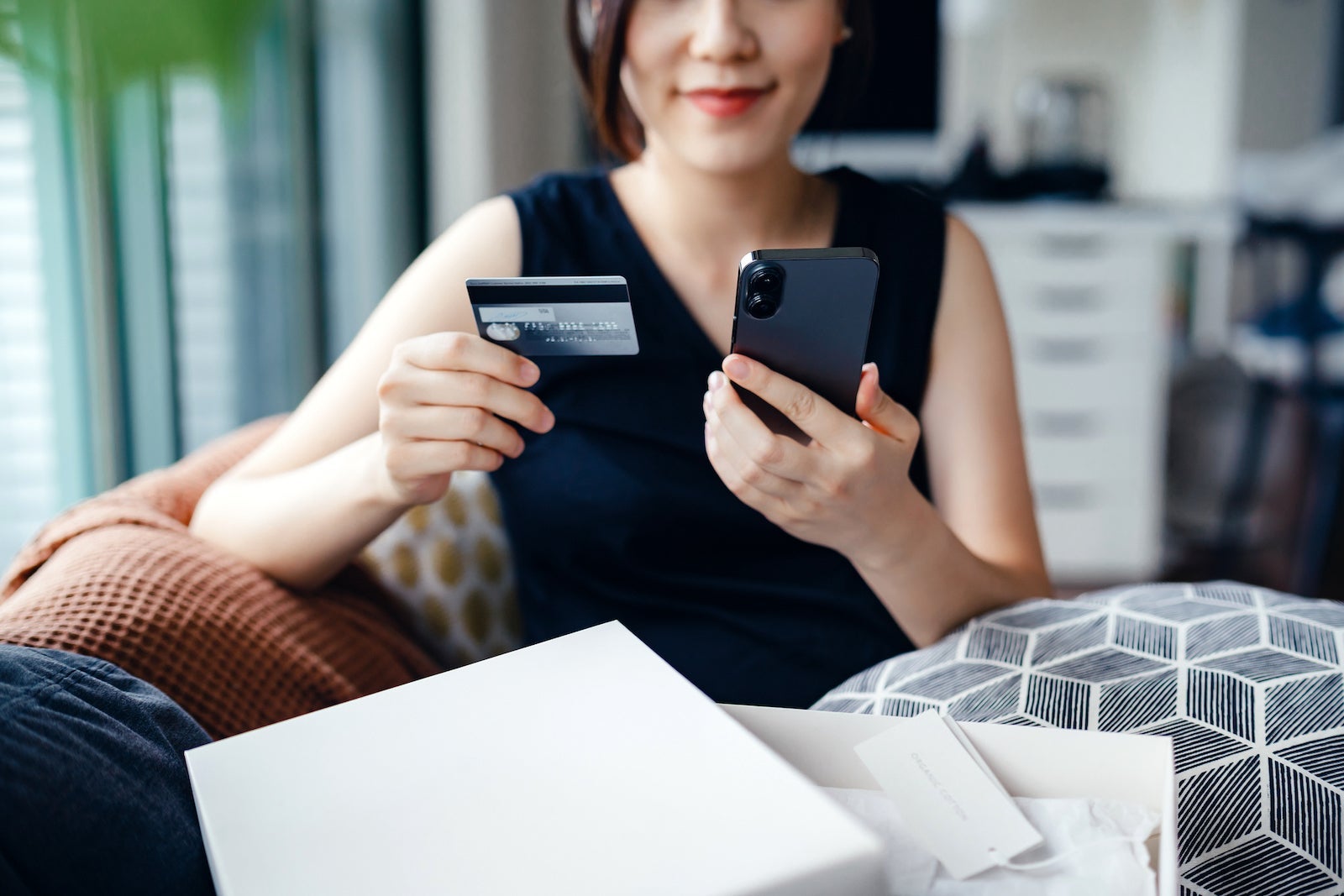 Beautiful smiling young Asian woman shopping online with smartphone and making payment online with credit card at home