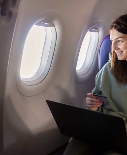 A young woman looks out a plane window while holding a credit card and a laptop