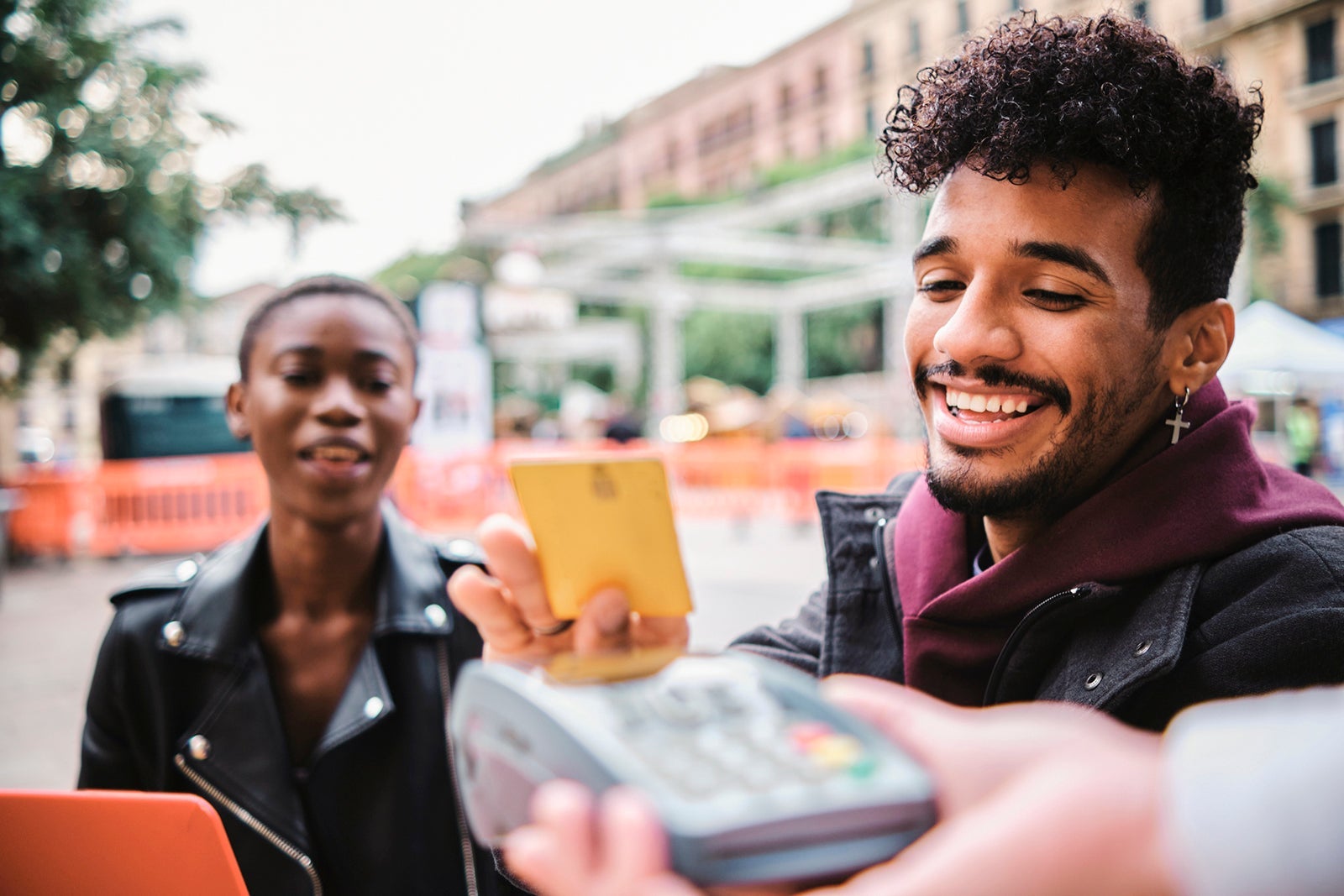 Smiling man paying through card whiles sitting with friend at sidewalk cafe