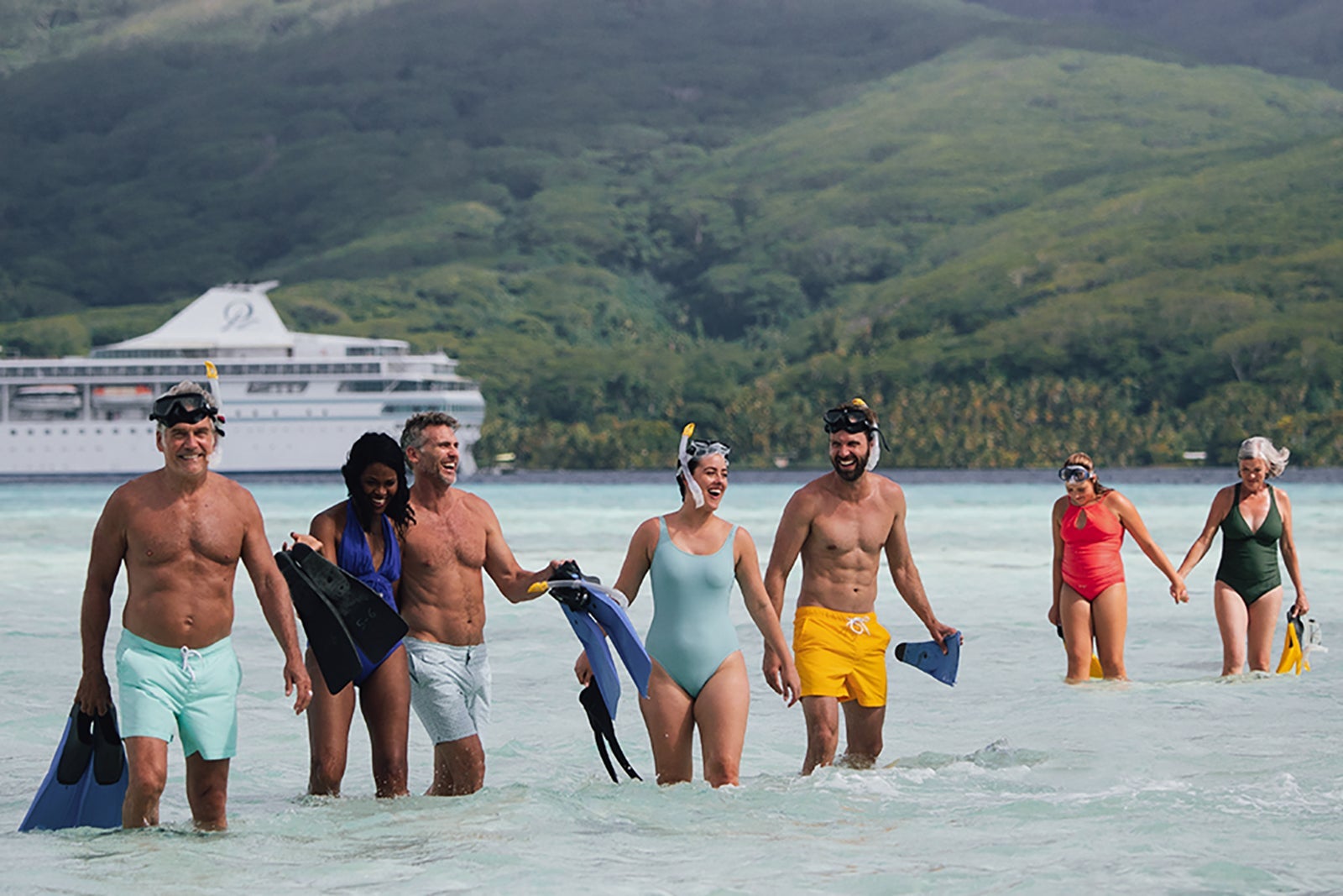 A group of snorkelers wades out of the water carrying their gear while their cruise ship waits in the background