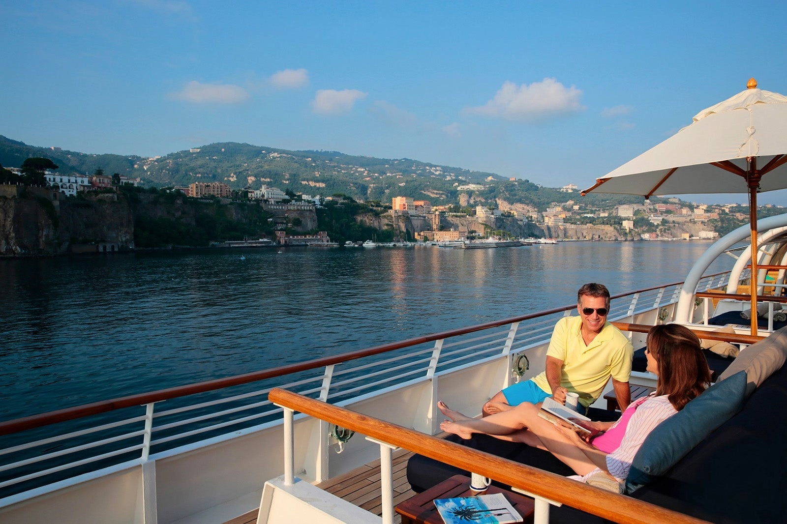 A couple sits on the outer deck of a cruise ship, smiling and watching the scenery