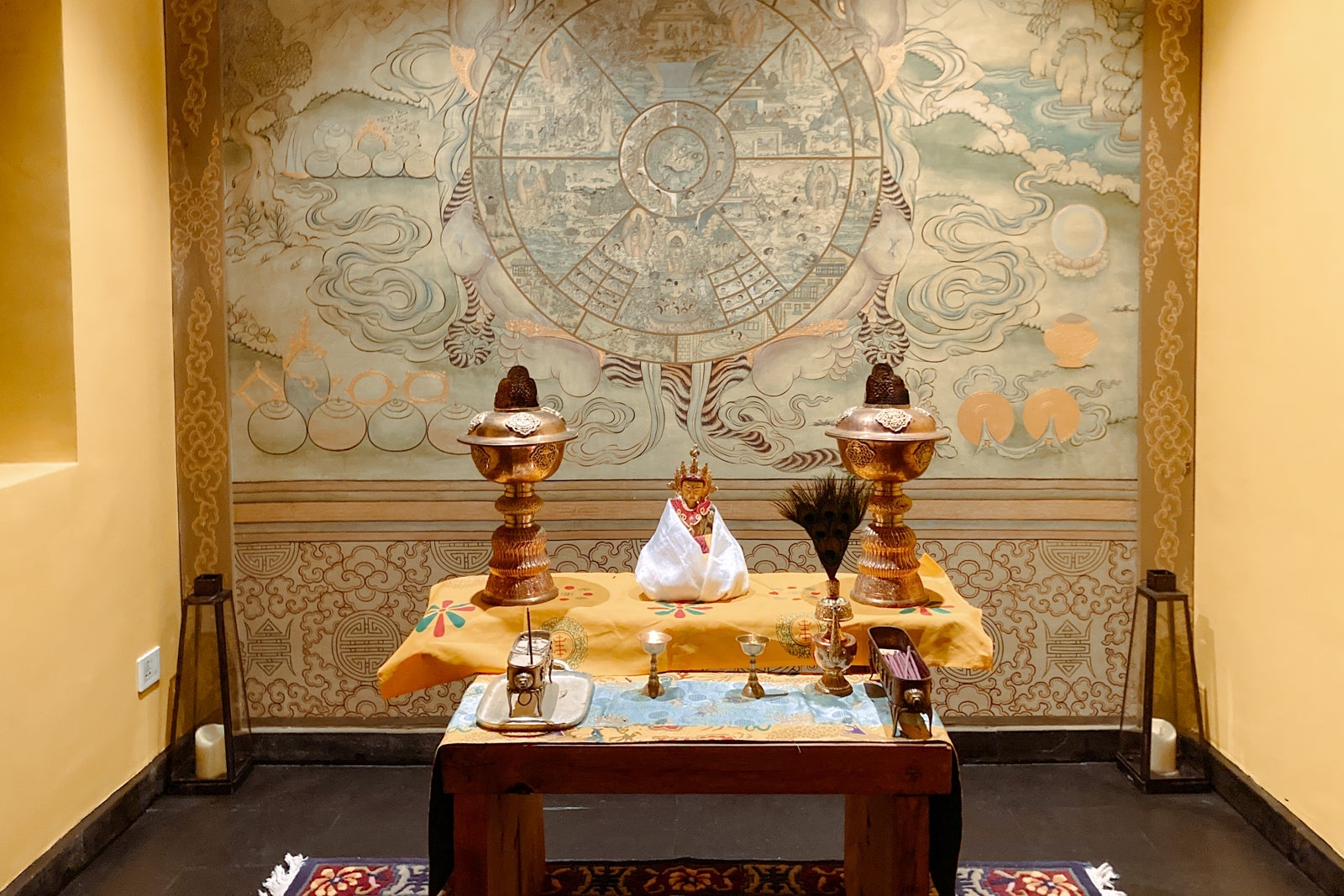 an altar with incense burning, shown in front of a wall covered with a traditional wheel of life Buddhist painting