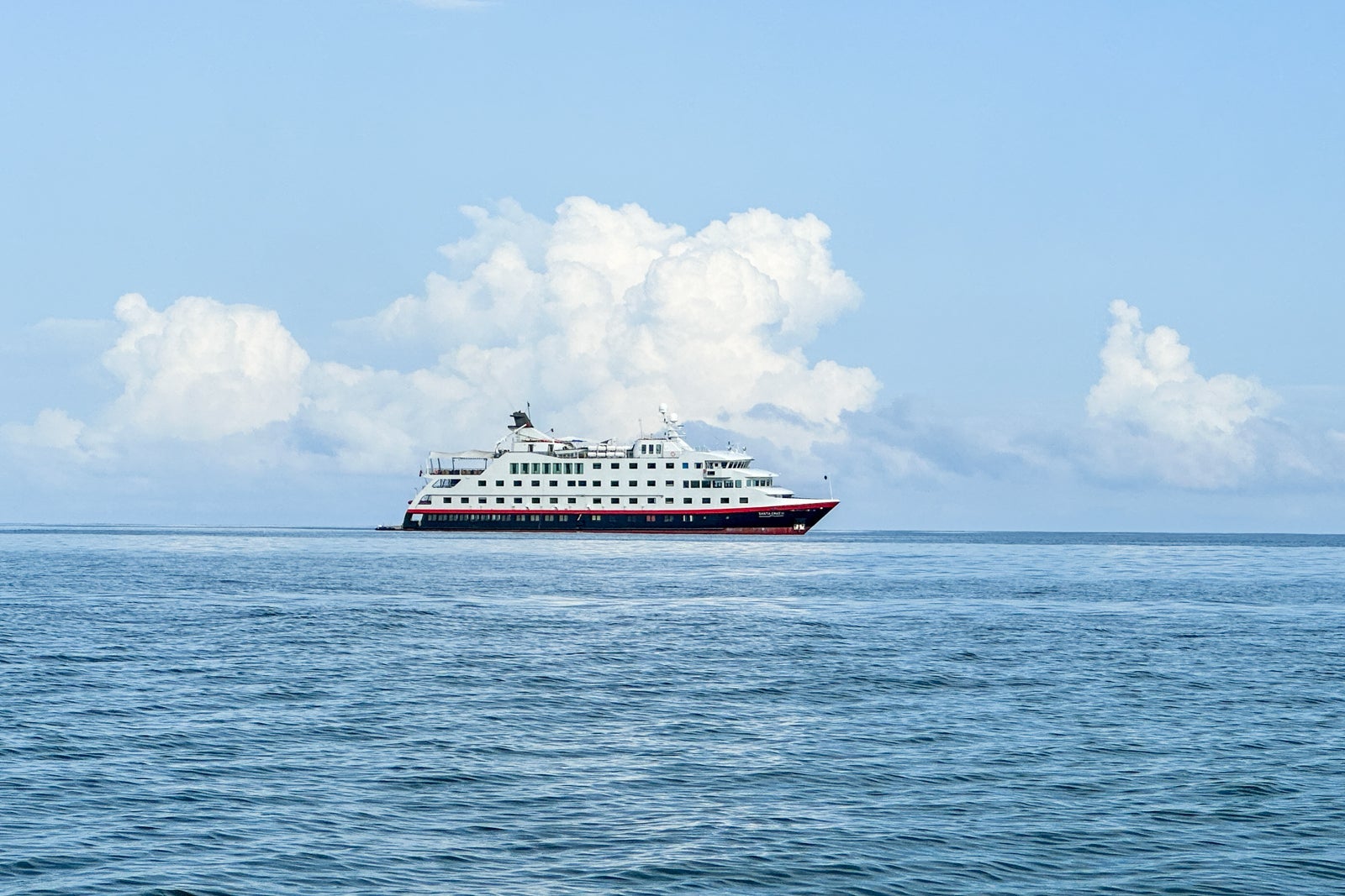 An expedition cruise ship from far way floating on the water with a cloudy sky behind it
