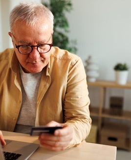 a senior man sits at a kitchen table, holding a credit card while online shopping