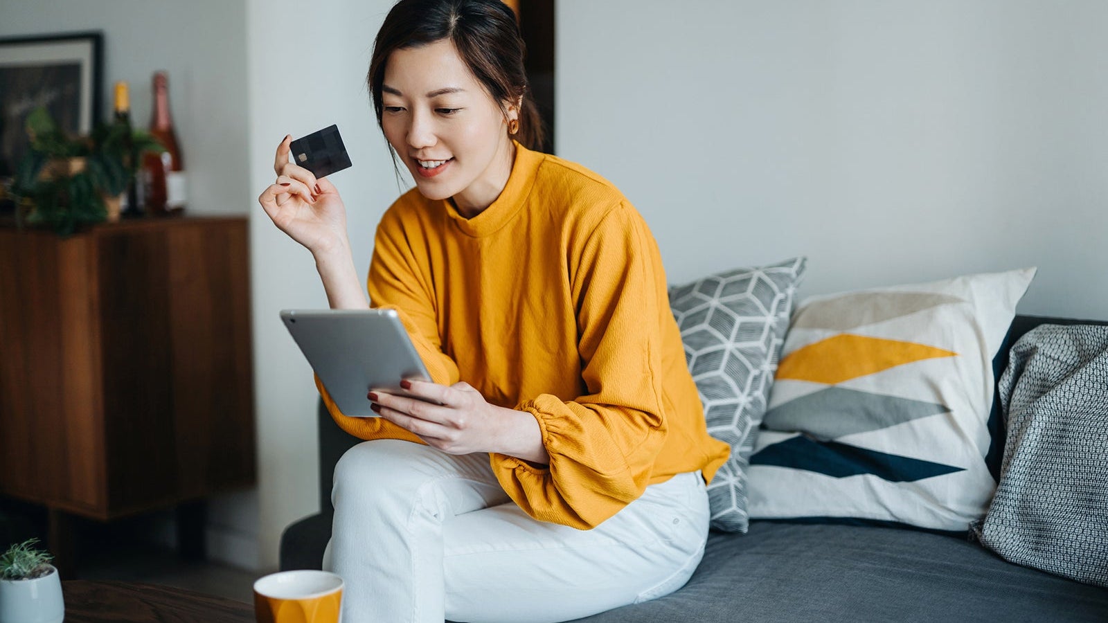 Beautiful smiling young Asian woman with credit card managing personal banking and finance with online banking mobile app on digital tablet at cozy home. Wealth management. Digital banking habits. Smart banking with technology