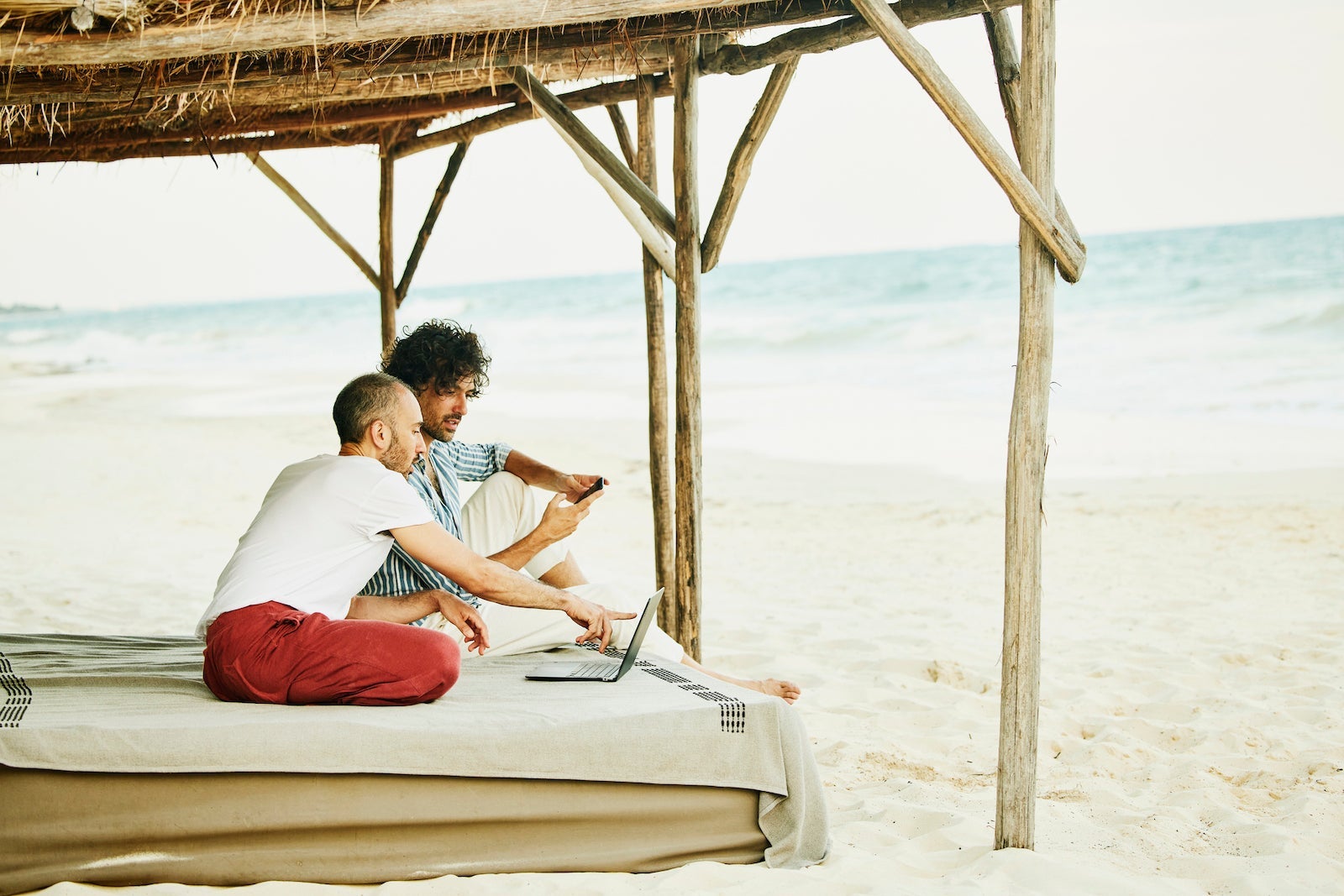 a gay couple sits at a beachfront cabana while using a laptop