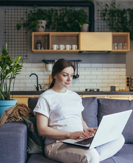 a woman sits on her sofa while using her laptop; a kitchen is visible in the background