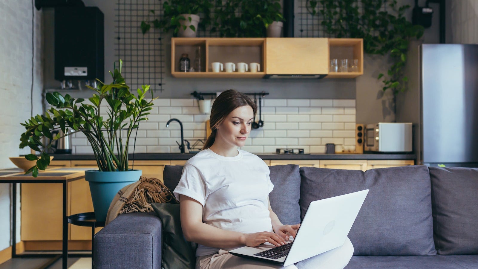 Maternity leave. Pregnant beautiful woman sitting on sofa at home with laptop in home clothes, ordering baby goods online, choosing, buying clothes and toys for baby.