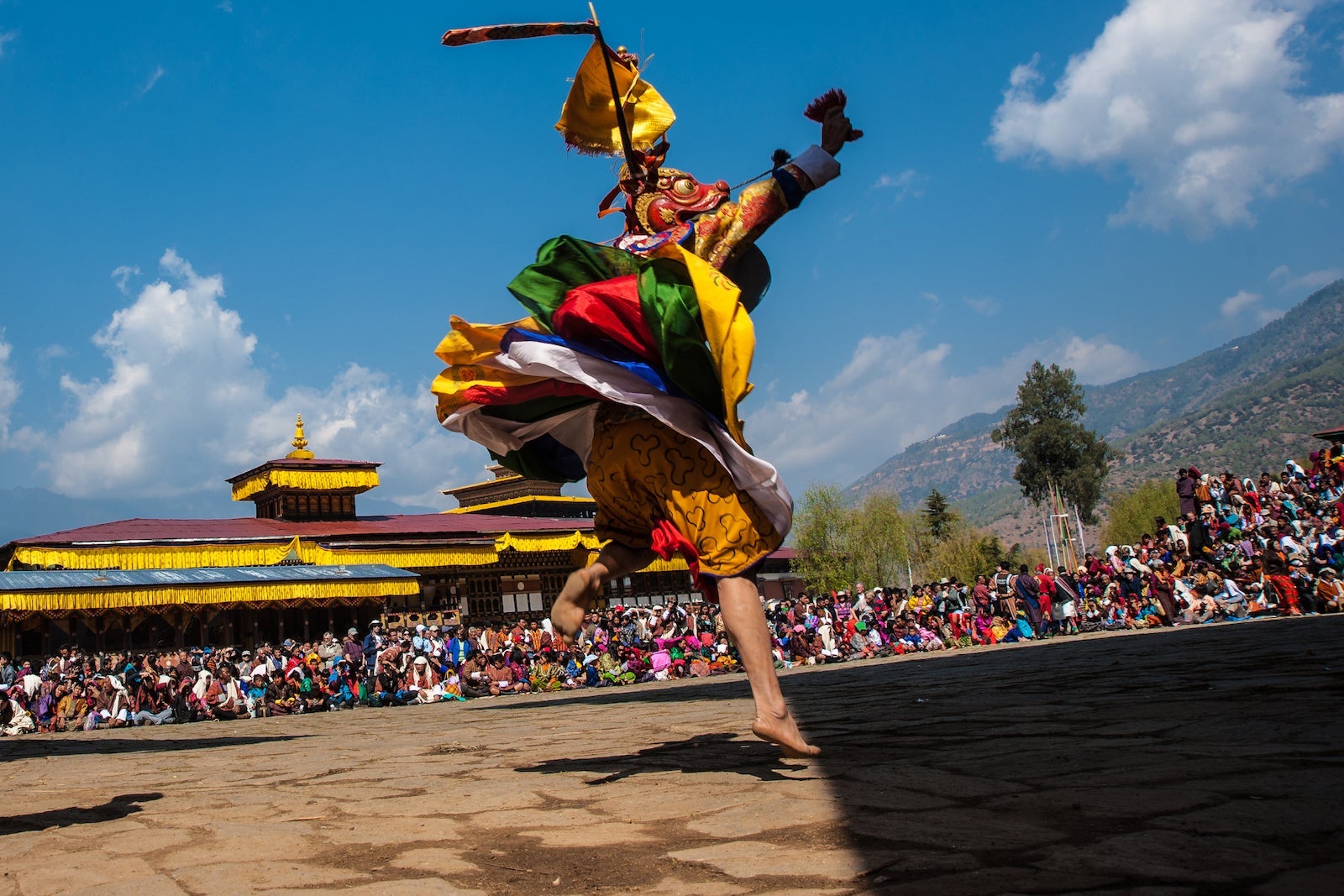 A monk in an elaborate costume performs a dance during a religious festival with many spectators in Bhutan