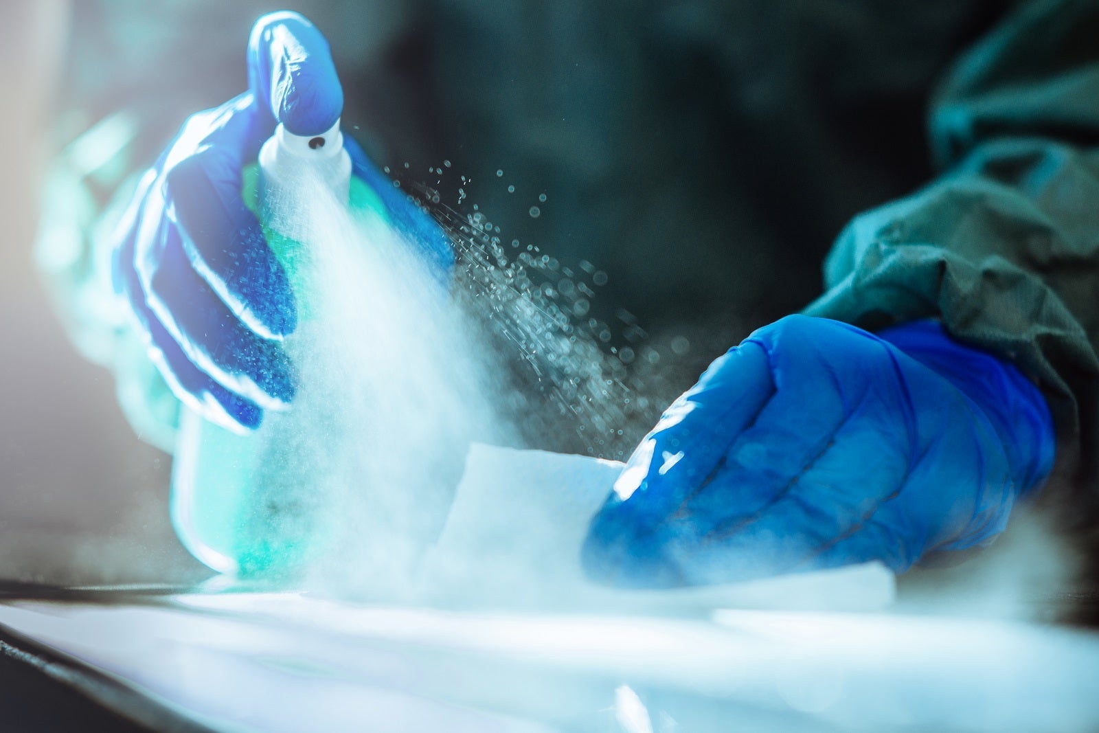 Two hands wearing rubber gloves -- one holding a spray bottle of sanitizer and the other holding a cleaning rag to wipe down a table