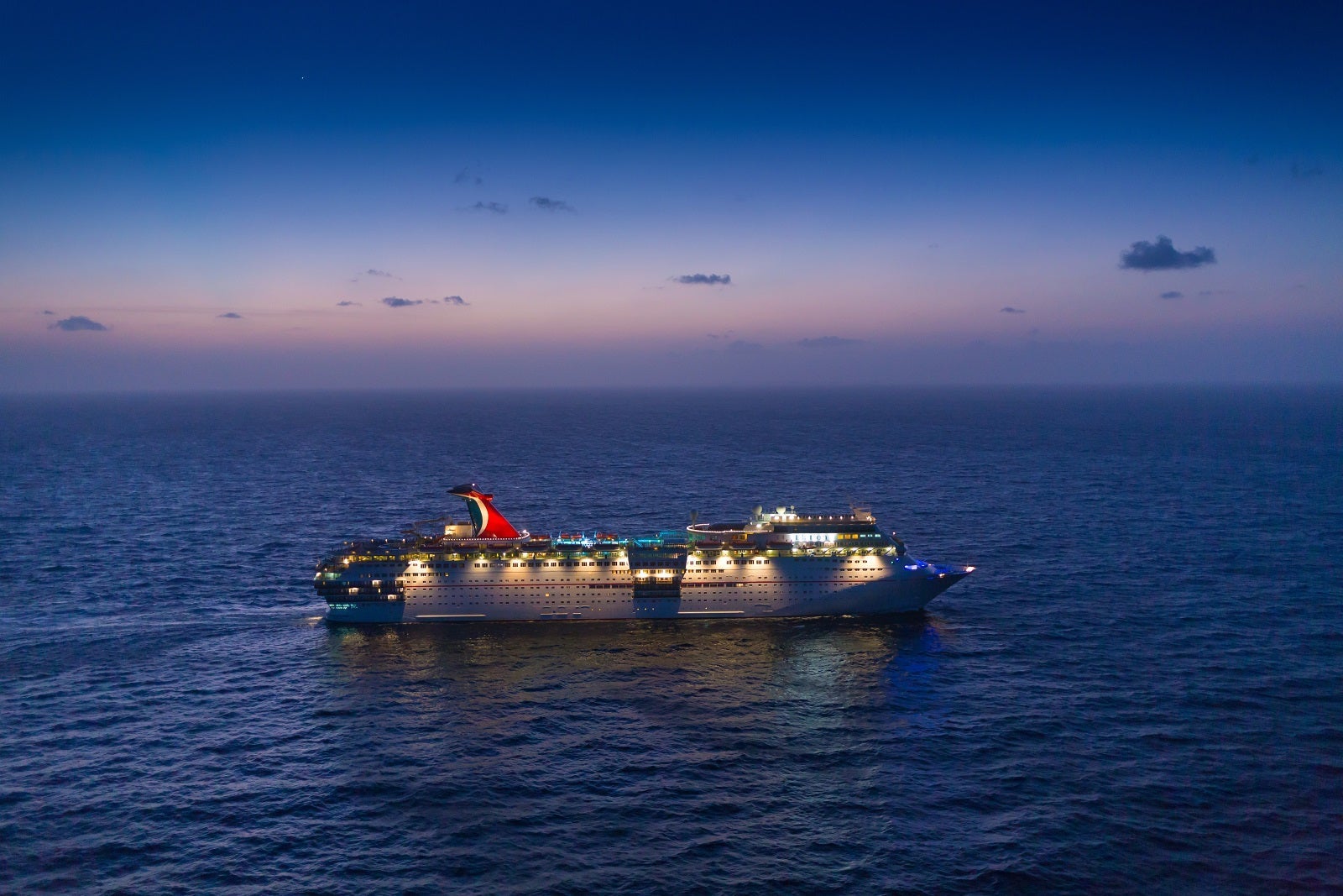 A side view of a cruise ship lit up at dusk while anchored at sea under a blue-purple sky