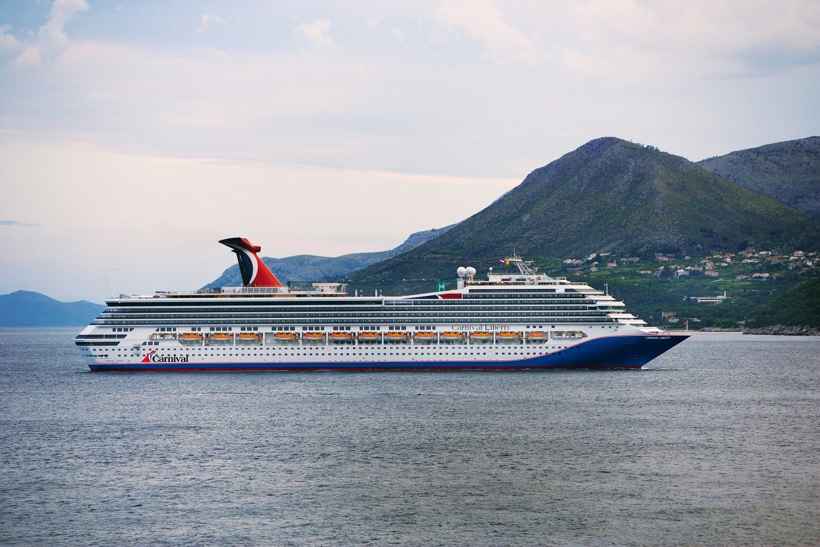 A cruise ship with a blue hull and a red, white and blue funnel sailing in front of a mountain