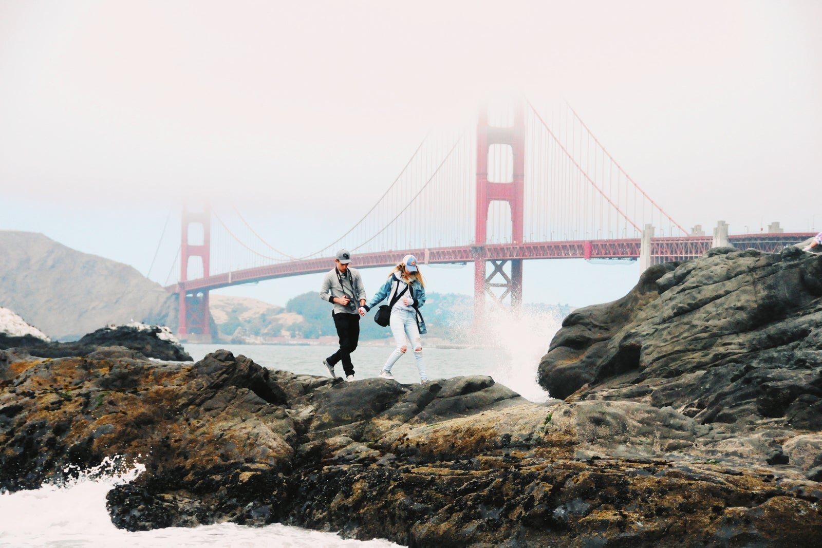 A couple walking along rocks in San Francisco's bay near the Golden Gate Bridge