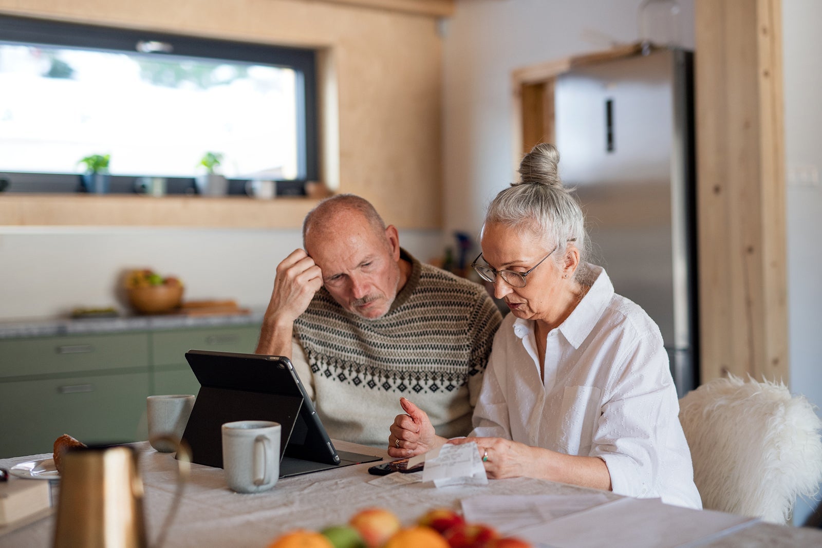 Senior couple sitting at the kitchen table looking at digital tablet and recalculating their expenses.