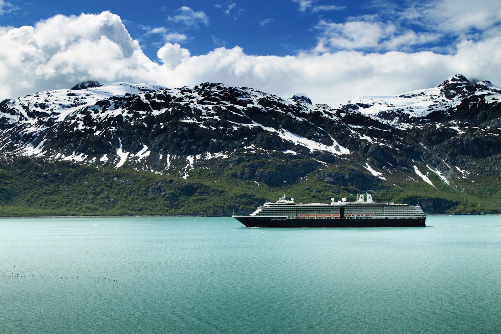 A Holland America cruise ship anchored on green water near a snow-covered mountain