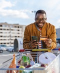 Portrait of a happy young African-American man pulling a shopping cart and using smart phone and credit card