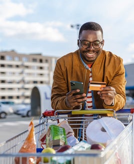 Portrait of a happy young African-American man pulling a shopping cart and using smart phone and credit card