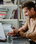 Smiling Young Man Wearing a Brown Turtleneck Sweater Paying his Bills on his Laptop Computer (Side View)