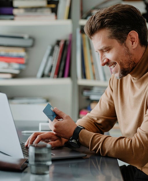 Smiling Young Man Wearing a Brown Turtleneck Sweater Paying his Bills on his Laptop Computer (Side View)