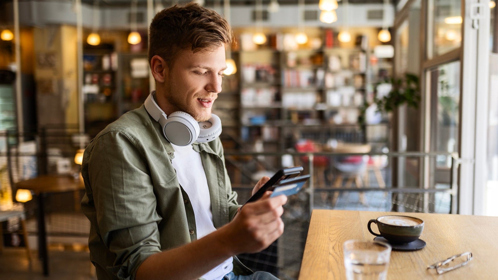 Happy man sitting and drinking coffee at cafe using his phone with credit card