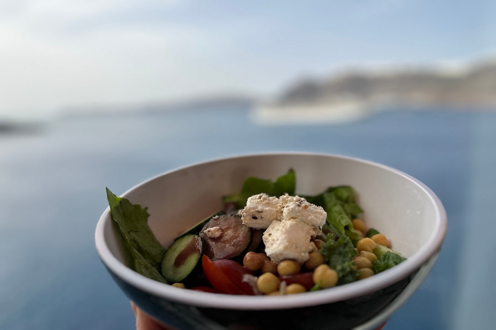 A bowl of salad in front of a window overlooking the ocean