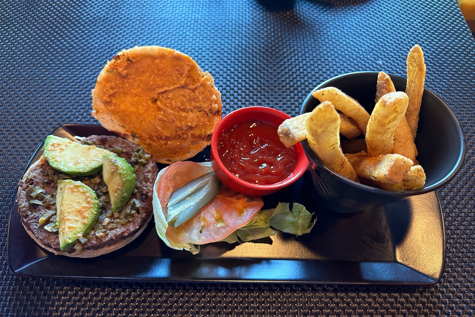 An open-faced veggie burger with avocado, lettuce, tomato, fries and ketchup on a black rectangular plate
