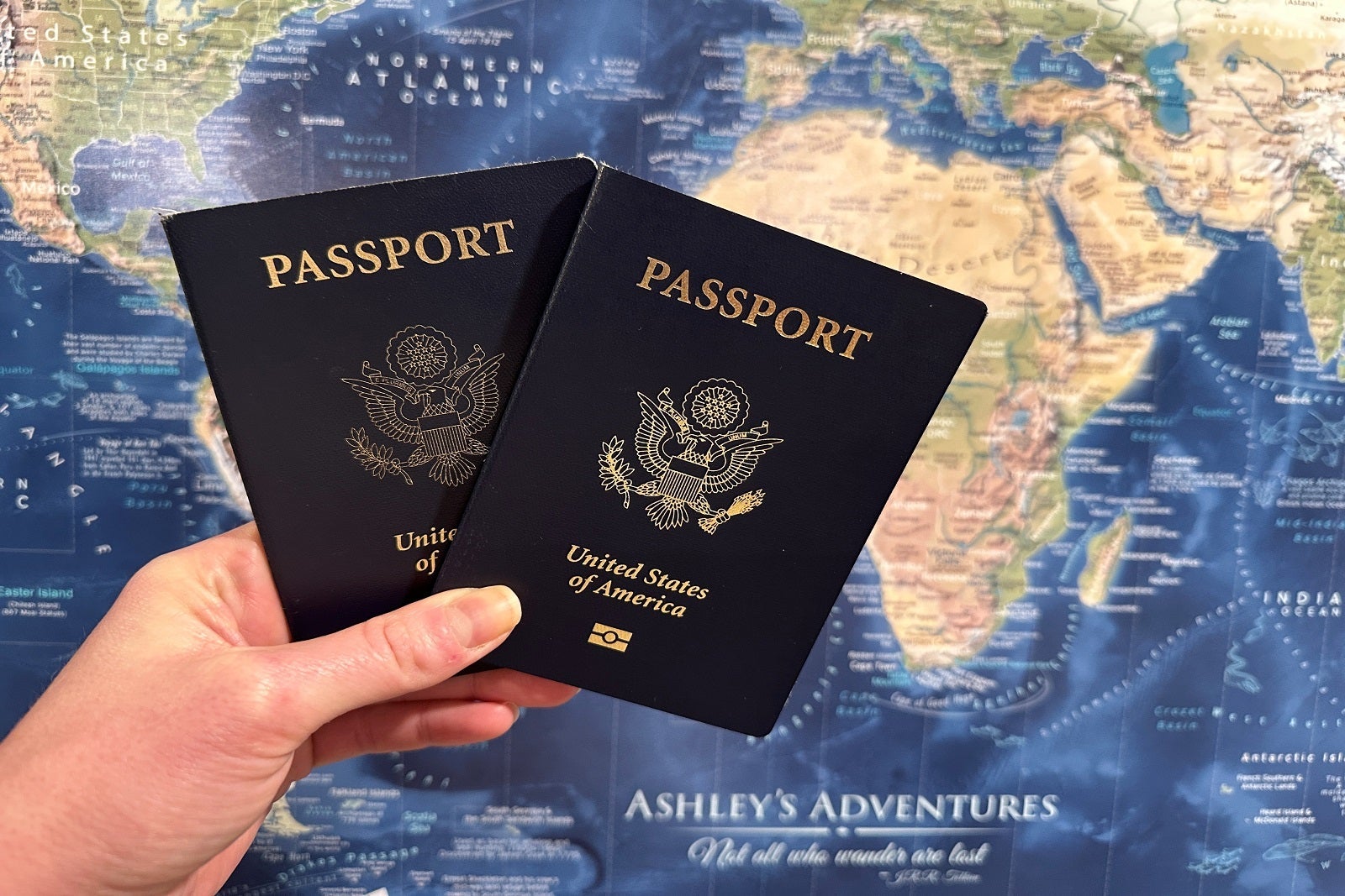 A hand holds two U.S. passports in front of a wall map.