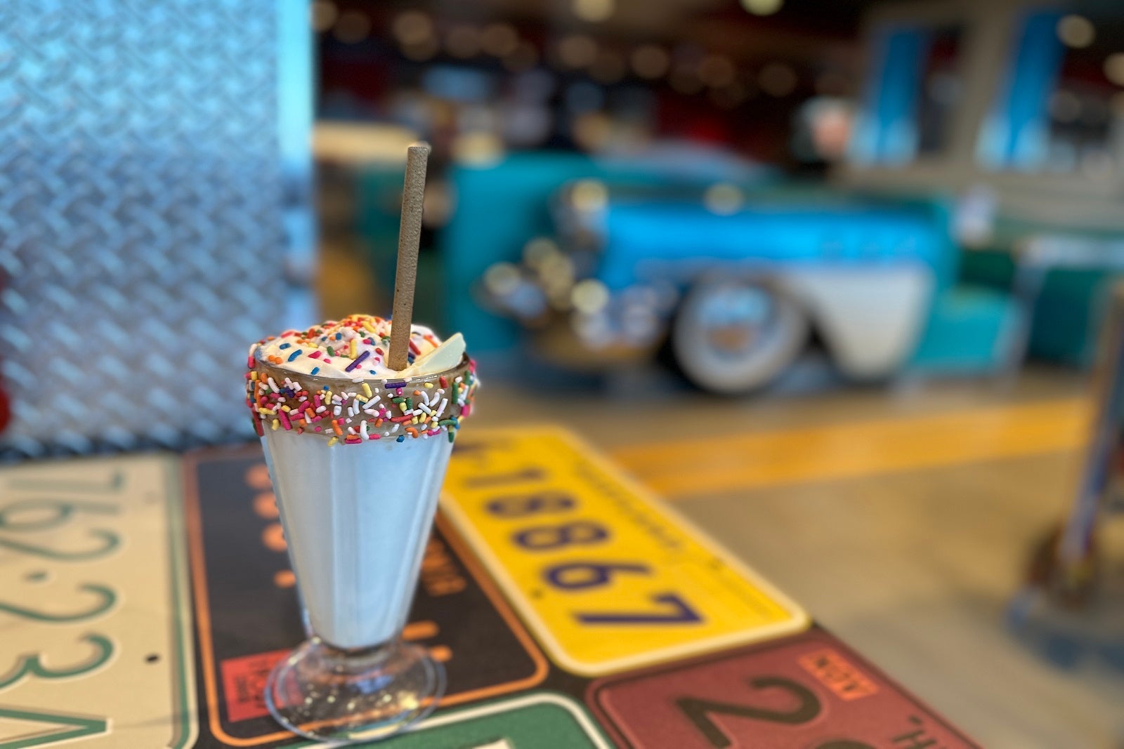A vanilla milkshake with sprinkles and a straw in a tall glass on a table decorated with license plates and a vintage car in the background