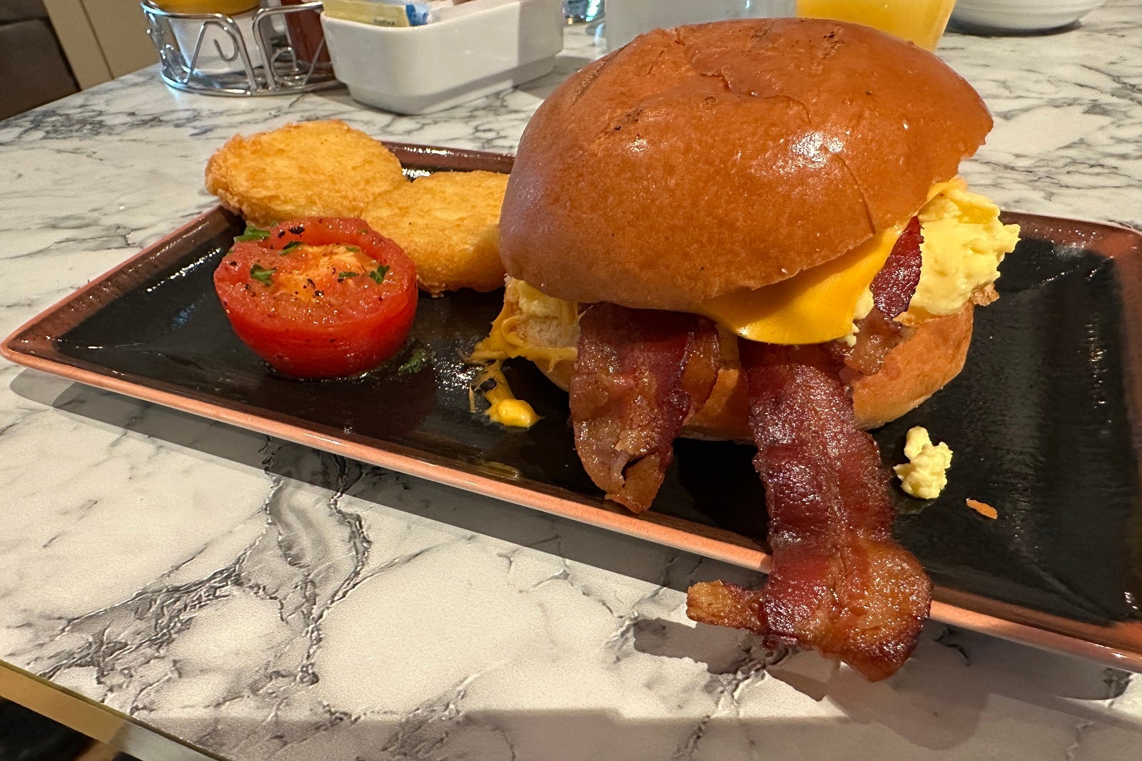 A breakfast sandwich with a tomato and hash browns on a black rectangular plate