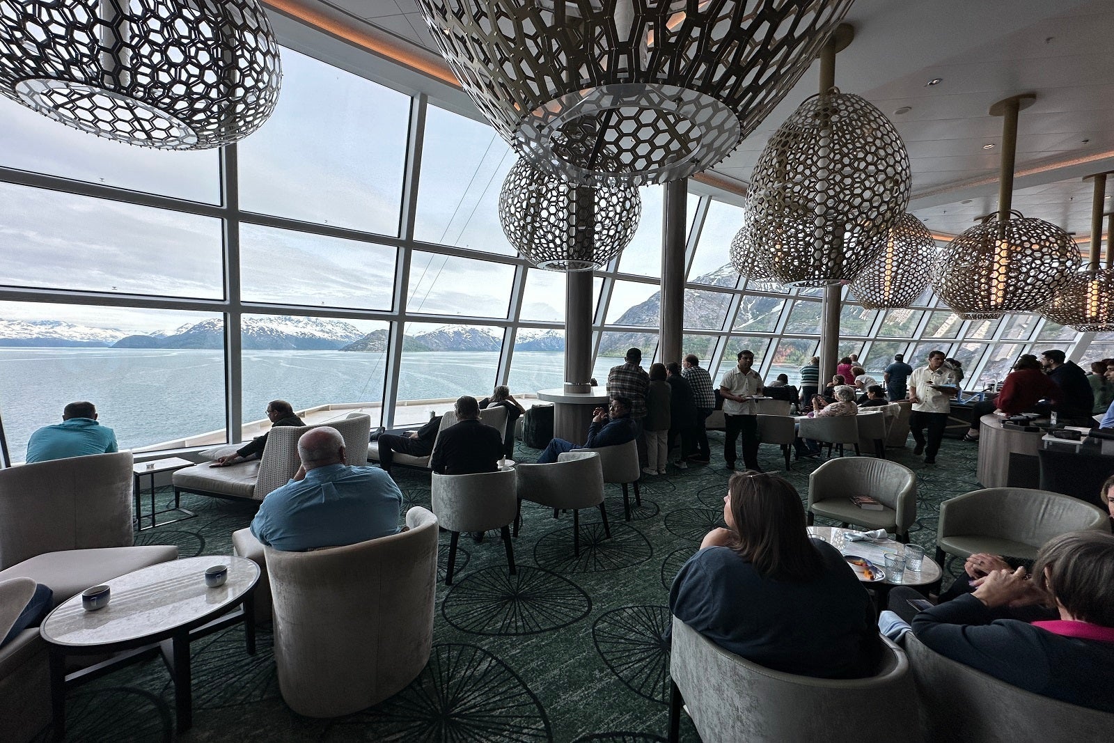 A cruise ship lounge with passengers staring out the floor-to-ceiling windows at glaciers