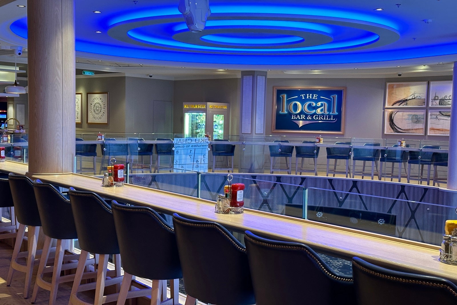 A row of bar seating overlooking a cruise ship atrium with a sign in the background that says "The Local"
