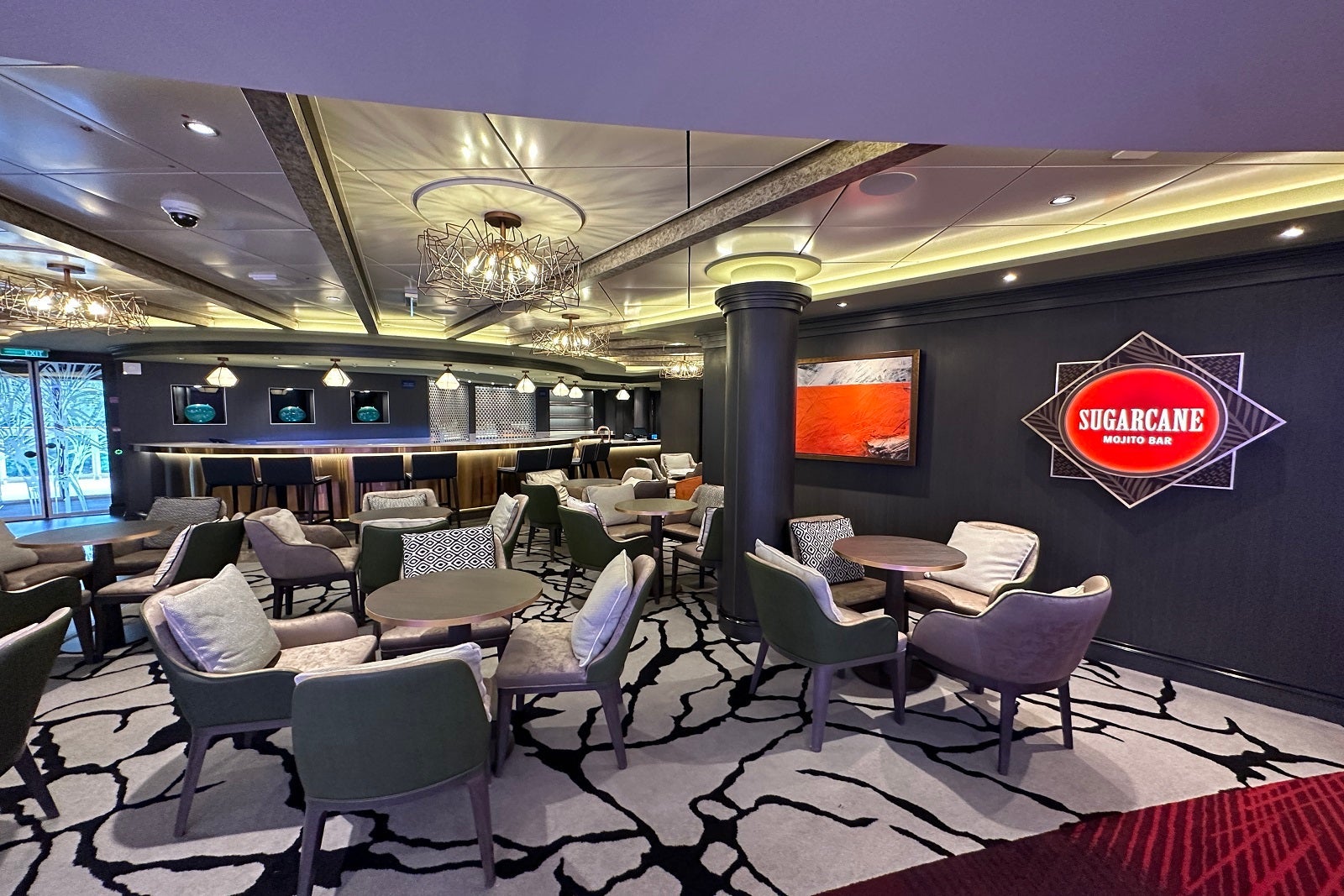 A cruise ship bar with a chandelier, gray carpet and chairs, tables and a red sign that says "Sugarcane Mojito Bar"