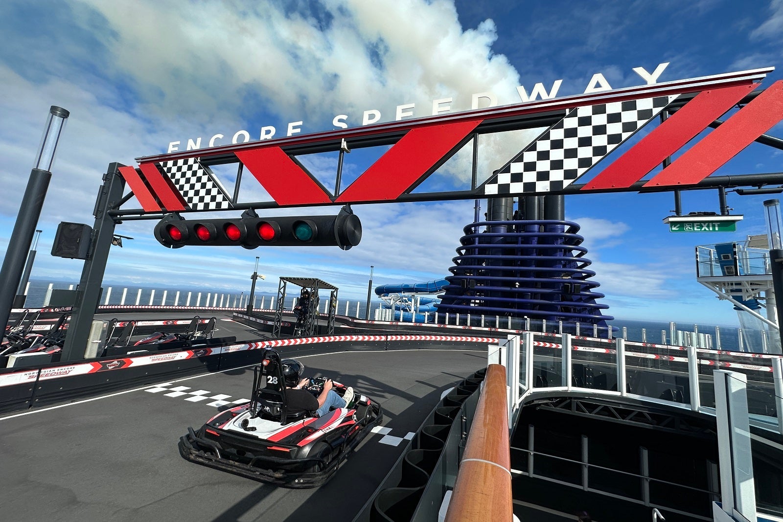 A passenger zooms by on a cruise ship go-kart and passes under a checkpoint that says "Encore Speedway"