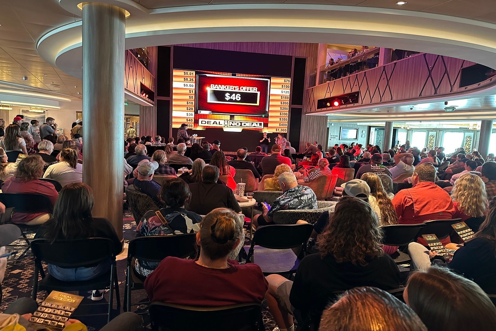 Passengers pack seats in a cruise ship atrium to watch a game of "Deal or No Deal" on a large screen