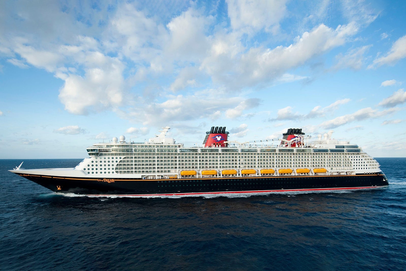 A cruise ship with a black hull and two red funnels sailing on dark blue water with a blue sky and clouds overhead