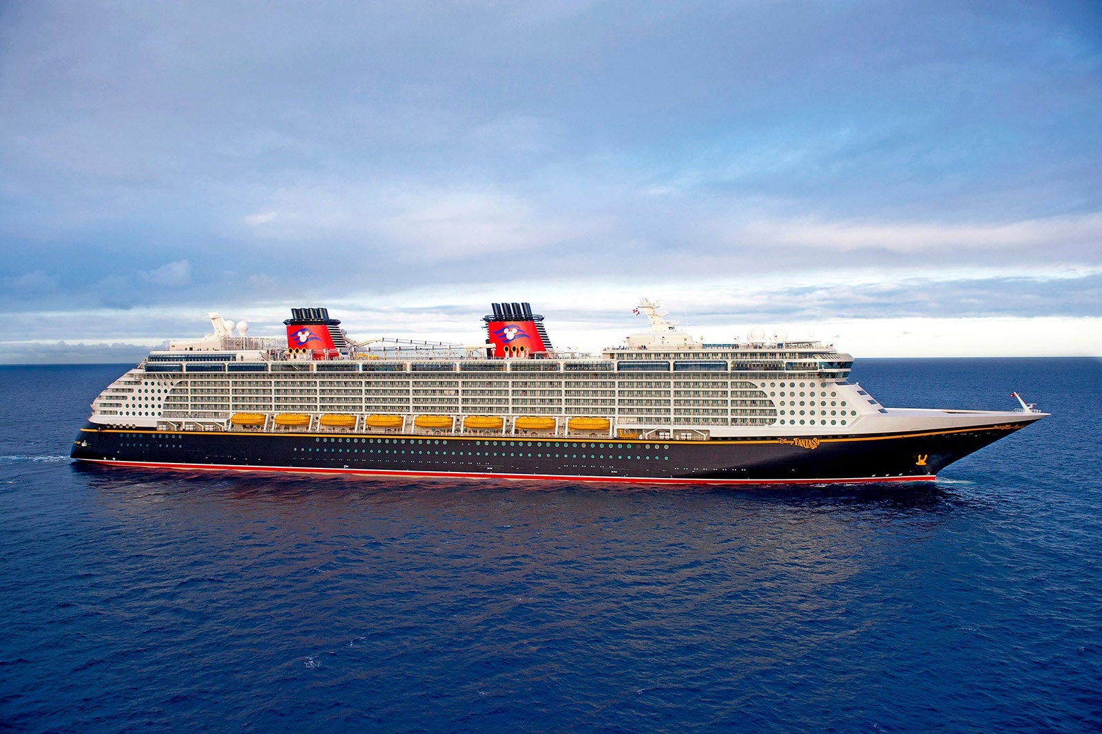 A Disney cruise ship with a black hull and red smokestacks anchored offshore on blue water with a blue sky overhead