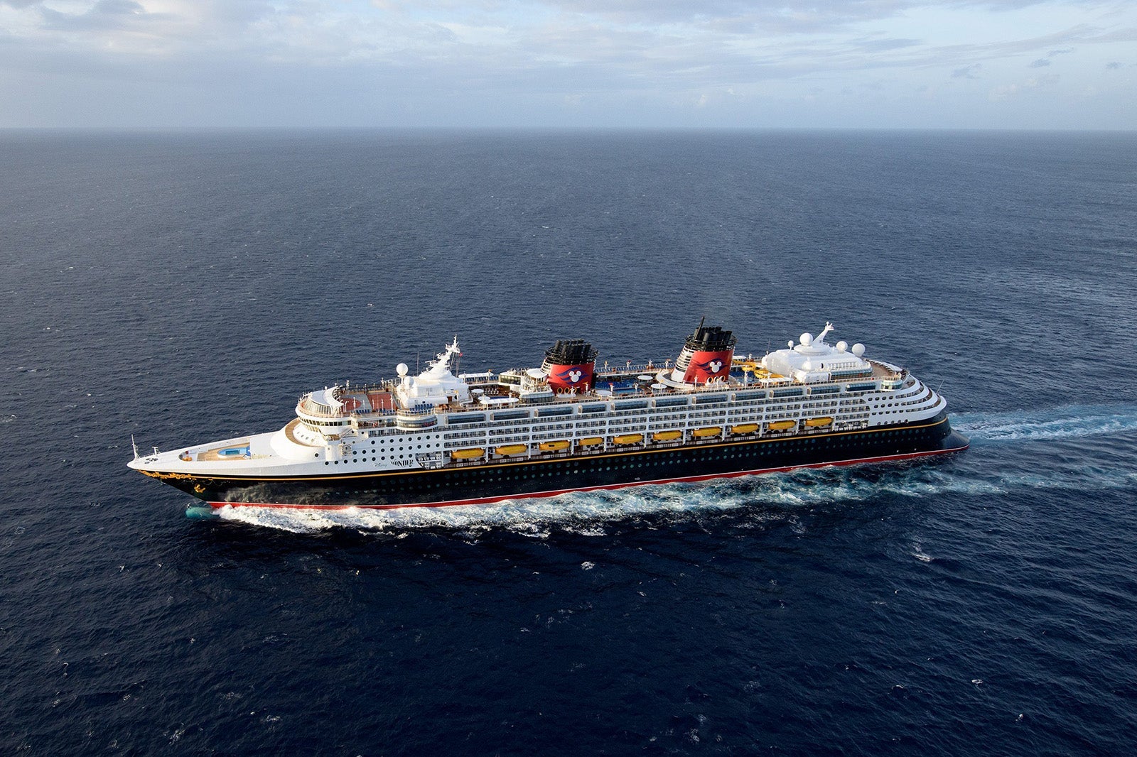 A cruise ship with a black hull and red smokestacks sailing on dark blue water with blue sky overhead