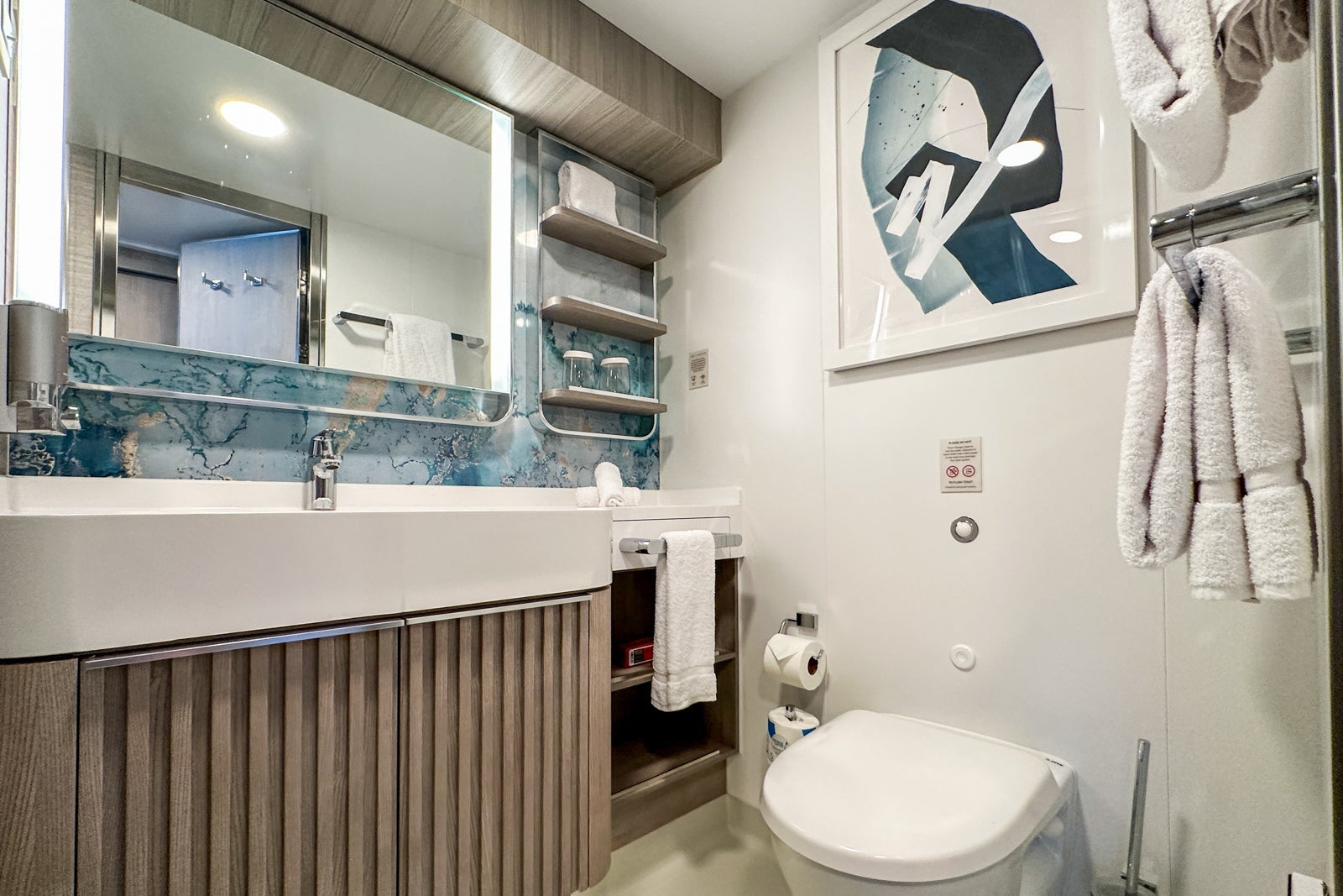 A cruise ship bathroom with a vanity, mirror, toilet, shelving and artwork on the wall