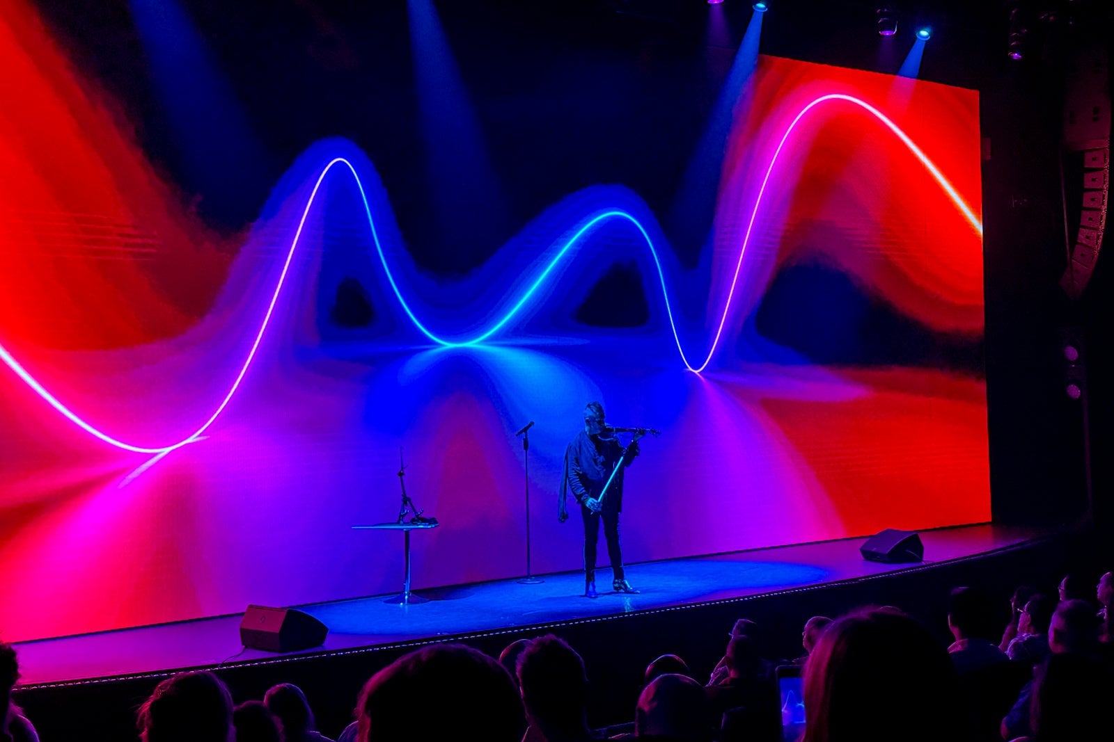 A cruise ship performer playing a violin on the ship's stage in the main theater with wavy, colorful graphics on a screen behind him