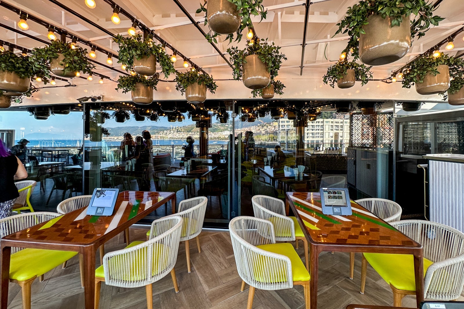 An outdoor dining area on a cruise ship with tables, chairs and plants hanging overhead against a glass window