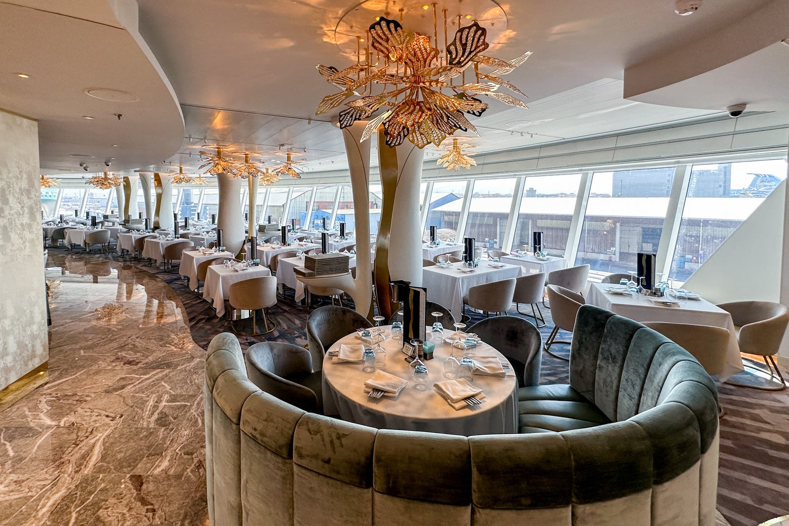 A cruise ship main dining room with banquette seating, tables and chairs with posts that look like gold trees
