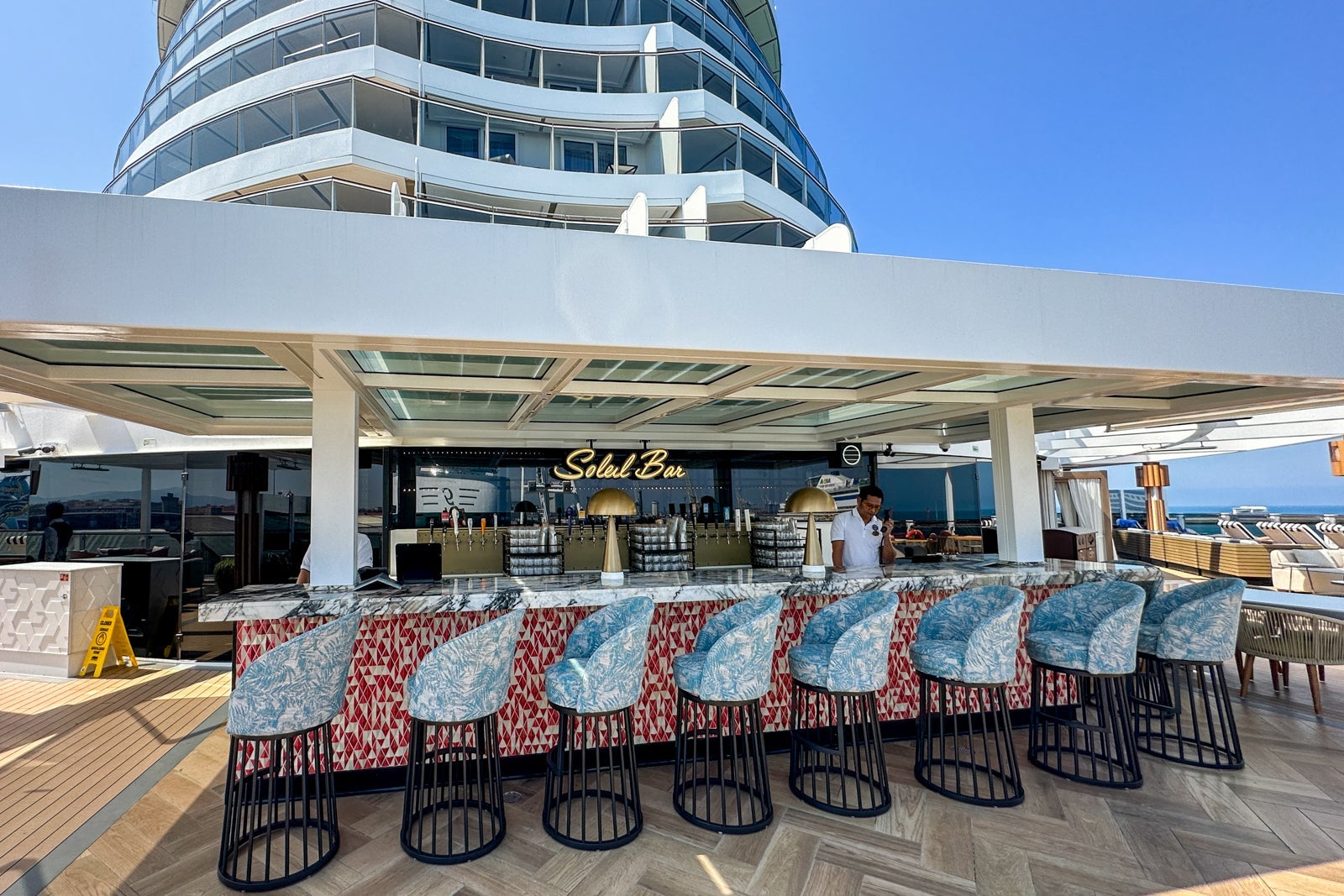 An outdoor bar with a red front and silver and black chairs on a cruise ship